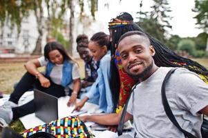 gruppo di cinque studenti universitari africani che trascorrono del tempo insieme nel campus nel cortile dell'università. amici afro neri seduti sull'erba e che studiano con i laptop. foto