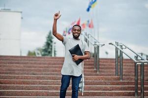 studente africano maschio posato con zaino e articoli per la scuola nel cortile dell'università, contro le bandiere di diversi paesi. mostra il dito in su. foto