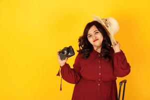 donna attraente del viaggiatore del sud asiatico in abito rosso intenso, cappello posato in studio su sfondo giallo con valigia e vecchia macchina fotografica vintage. foto