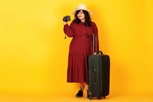 donna attraente del viaggiatore del sud asiatico in abito rosso intenso, cappello posato in studio su sfondo giallo con valigia e vecchia macchina fotografica vintage. foto