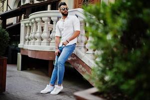 elegante uomo arabo alto modello in camicia bianca, jeans e occhiali da sole posato in una strada della città. barba attraente ragazzo arabo. foto