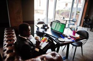 uomo d'affari afroamericano indossare su abito nero e occhiali seduto in ufficio con laptop schermo verde e lavorare, leggere riviste e bere caffè. foto