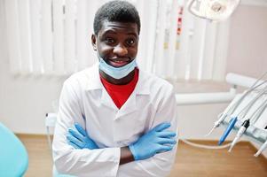 medico maschio afroamericano in maschera con le braccia incrociate seduto alla poltrona del dentista in clinica dentale. foto