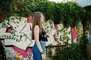 elegante ragazza hipster casual con berretto, occhiali e jeans indossano, ascoltando musica dalle cuffie del telefono cellulare contro il grande muro di graffiti. foto