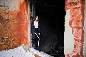 modello bruna alla moda gambe lunghe in lungo mantello nero poste all'aperto al giorno d'inverno contro il vecchio muro di lerciume. foto