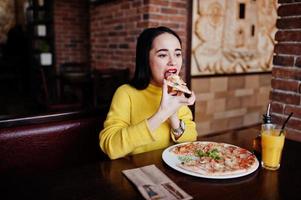 divertente ragazza bruna in maglione giallo che mangia pizza al ristorante. foto