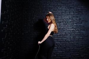 bella ragazza bruna indossare su nero, seduto e in posa su una sedia in studio contro il muro di mattoni scuri. ritratto del modello in studio. foto