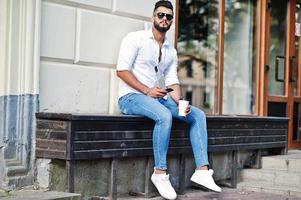 elegante uomo arabo alto modello in camicia bianca, jeans e occhiali da sole posato in una strada della città e tenere il cellulare a portata di mano. ragazzo arabo con la barba con una tazza di caffè seduto e riposando. foto