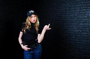 agente femminile dell'FBI in berretto e con la pistola in studio contro un muro di mattoni scuri. foto