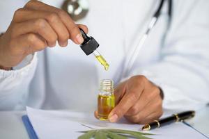 mani del medico che tengono un contagocce con un prodotto petrolifero sullo sfondo delle foglie e fiori di marijuana, olio di canapa cbd. foto