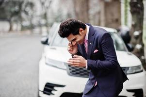 elegante modello di uomo macho indiano su abito e cravatta rosa in posa contro un'auto bianca e parlando al telefono. foto