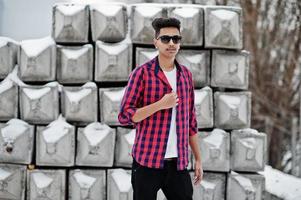 casual giovane indiano in camicia a scacchi e occhiali da sole in posa contro blocchi di pietra. foto
