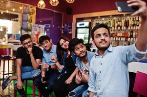 un gruppo di amici asiatici alla moda indossa jeans seduti su sedie contro il bar del club e fa selfie insieme. foto