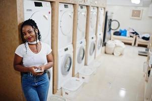 allegra donna afroamericana vicino alla lavatrice ascoltando musica con gli auricolari dal telefono cellulare nella lavanderia self-service. foto