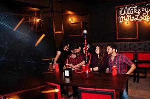 gruppo di amici indiani che si divertono e riposano in discoteca, bevono cocktail e fumano narghilè, guardando i telefoni cellulari. foto