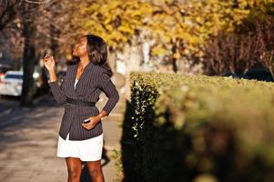 successo elegante donna afroamericana in giacca e gonna poste in una soleggiata giornata autunnale in strada. foto