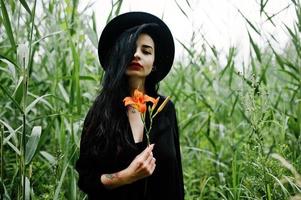 ragazza sensuale tutta in nero, labbra rosse e cappello. goth donna drammatica tenere fiore di giglio arancione su canna comune. foto