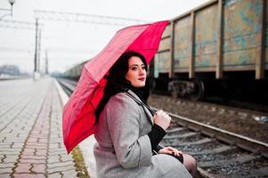 ragazza bruna in cappotto grigio con ombrello rosso nella stazione ferroviaria. foto