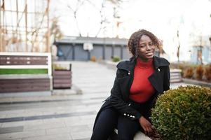 ritratto di una donna africana dai capelli ricci che indossa un cappotto nero alla moda e un dolcevita rosso in posa all'aperto. foto