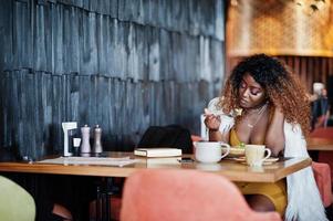 glamour donna afroamericana in abito giallo e mantello di lana bianco posato seduto al ristorante e mangiare insalata. foto