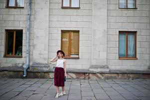 ragazza modello bionda alla moda e bella in elegante gonna in velluto di velluto rosso, camicetta bianca e cappello, in posa con telefono e auricolari. foto