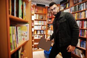 studente arabo alto e intelligente, indossa una giacca di jeans nera e occhiali da vista, in biblioteca con un libro a portata di mano. foto