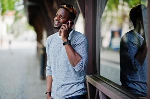 elegante ragazzo afroamericano su maglione grigio e occhiali da sole neri in posa per strada e parlando al telefono. ragazzo nero alla moda. foto