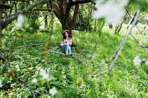 ritratto primaverile di una ragazza bruna con occhiali rosa e cappello al giardino di fiori verdi. foto