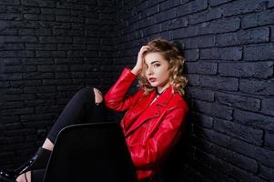 ritratto in studio di ragazza bionda in giacca di pelle rossa poste su una sedia contro un muro di mattoni. foto