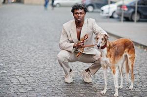 elegante uomo afro in abito beige vecchia scuola con cane borzoi russo. giovane maschio africano alla moda in giacca casual sul torso nudo. foto