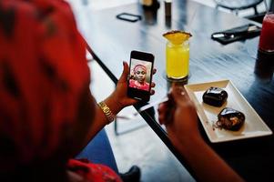 elegante donna africana in camicia rossa e cappello in posa in un bar al coperto, mangiare dolci al cioccolato e fare selfie al telefono cellulare. foto