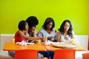 quattro giovani ragazze africane in un ristorante fast food dai colori vivaci che fanno foto di pizza nei loro telefoni cellulari.