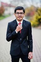 giovane indiano con gli occhiali, indossa un abito nero con cravatta rossa in posa all'aperto. foto