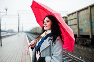 ragazza bruna in cappotto grigio con ombrello rosso nella stazione ferroviaria. foto