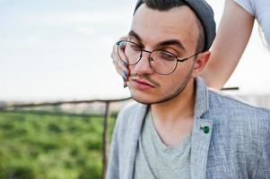 mano della ragazza sul viso del sognatore elegante uomo macho in abito grigio, cappello e occhiali posati sul tetto. foto