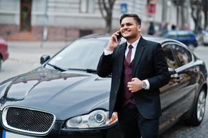 elegante uomo d'affari indiano in abbigliamento formale con il telefono cellulare in piedi contro un'auto nera d'affari sulla strada della città. parlare al cellulare. foto