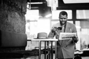 elegante uomo afroamericano modello in giacca grigia cravatta e cappello rosso bere caffè al bar e leggere i giornali. foto in bianco e nero.
