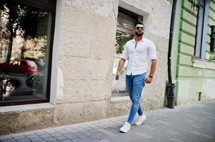 elegante uomo arabo alto modello in camicia bianca, jeans e occhiali da sole posato in una strada della città. barba attraente ragazzo arabo. foto