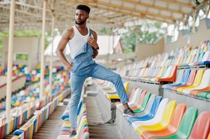 bell'uomo afroamericano in tuta di jeans posato su sedie colorate allo stadio. ritratto di uomo nero alla moda. foto