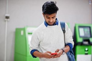 giovane uomo asiatico alla moda con il telefono cellulare e lo zaino contro la fila di bancomat verde. foto