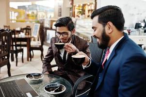 due uomini d'affari indiani in giacca e cravatta seduti in ufficio al bar, guardando il laptop e bevendo caffè. foto