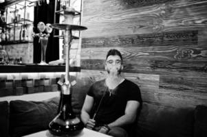 elegante barba uomo arabo in occhiali e t-shirt nera che fuma narghilè bar interno. modello arabo che riposa. foto