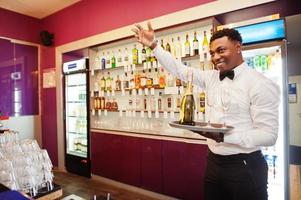 barista afroamericano al bar con champagne con bicchieri sul vassoio. foto