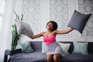 attraente donna afroamericana con capelli afro sulla gonna rosa in alto, posata in una stanza bianca. il modello nero sexy alla moda gioca con i cuscini. foto