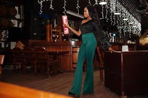 bellezza sottile modello afroamericano indossare su camicetta nera e pantaloni verdi gambe lunghe in posa contro decorazioni natalizie e tenere una confezione regalo per le celebrazioni del nuovo anno. foto