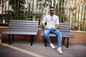 elegante uomo arabo alto modello in camicia bianca, jeans e occhiali da sole posato in una strada della città. barba attraente ragazzo arabo seduto su una panchina. foto