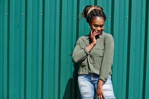 donna africana con i capelli dreadlocks, in pantaloncini di jeans poste contro il muro di acciaio verde. foto