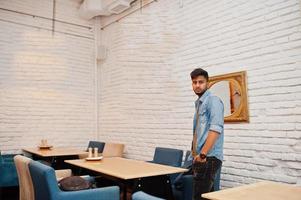 ritratto di bel successo barbuto del sud asiatico, giovane libero professionista indiano in camicia di jeans blu in piedi nella caffetteria e guardare lo specchio. foto