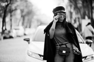 ragazza di moda afroamericana in cappotto, berretto da strillone e occhiali da sole poste in strada contro un'auto bianca da lavoro. foto