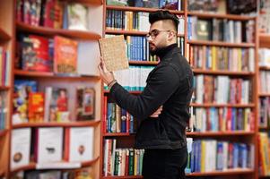 studente arabo alto e intelligente, indossa una giacca di jeans nera e occhiali da vista, in biblioteca con un libro a portata di mano. foto